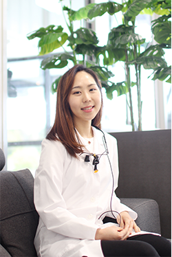 Portrait of Dr. Su-Young Jane Choi seated in dental office wearing white coat and loupes – Loving Dentistry